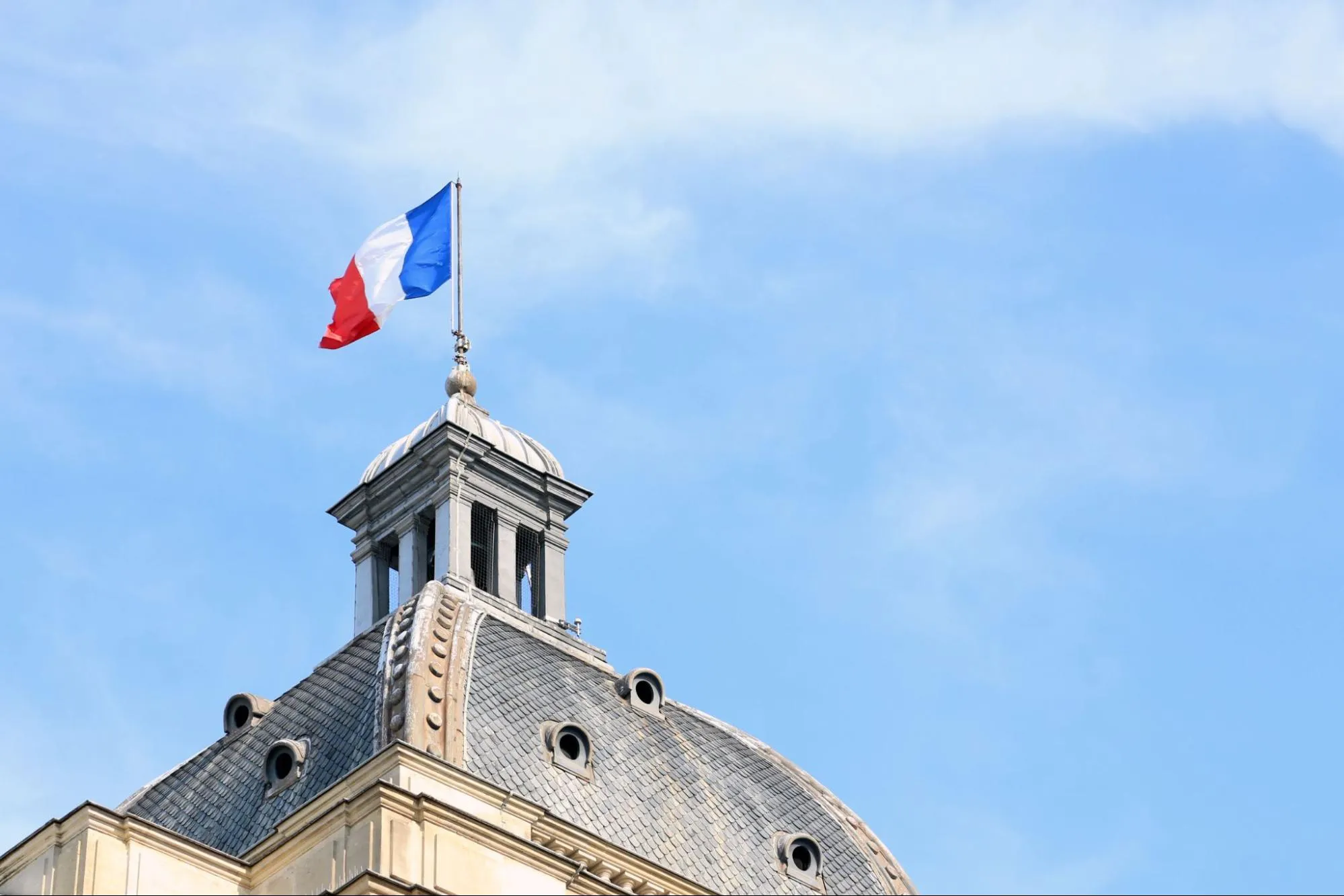 The French flag flying from a mast above the slate dome of a public building against a blue sky