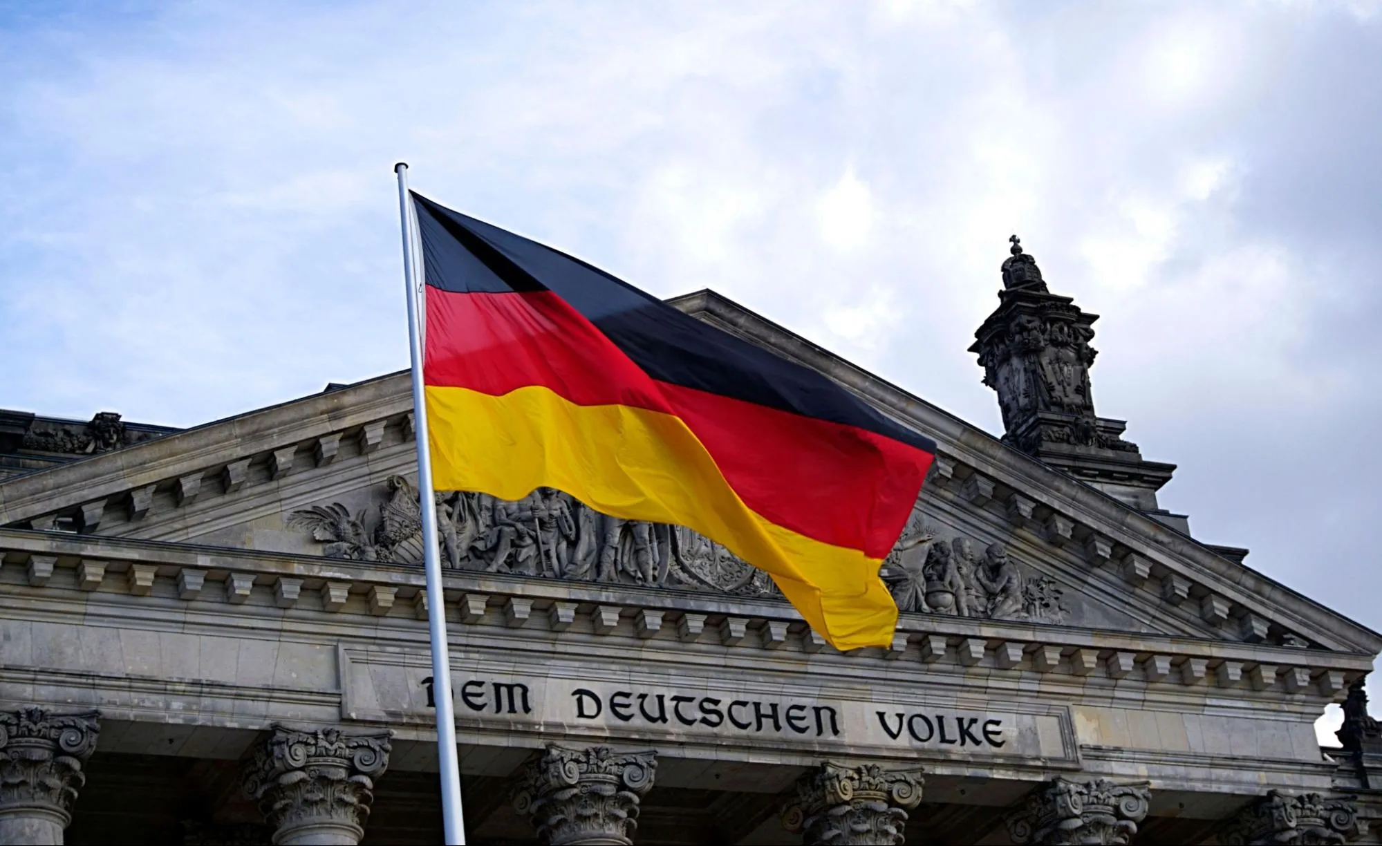 The German flag flying in front of the Reichstag building in Berlin, with the inscription DEM DEUTSCHEN VOLKE visible on the architrave