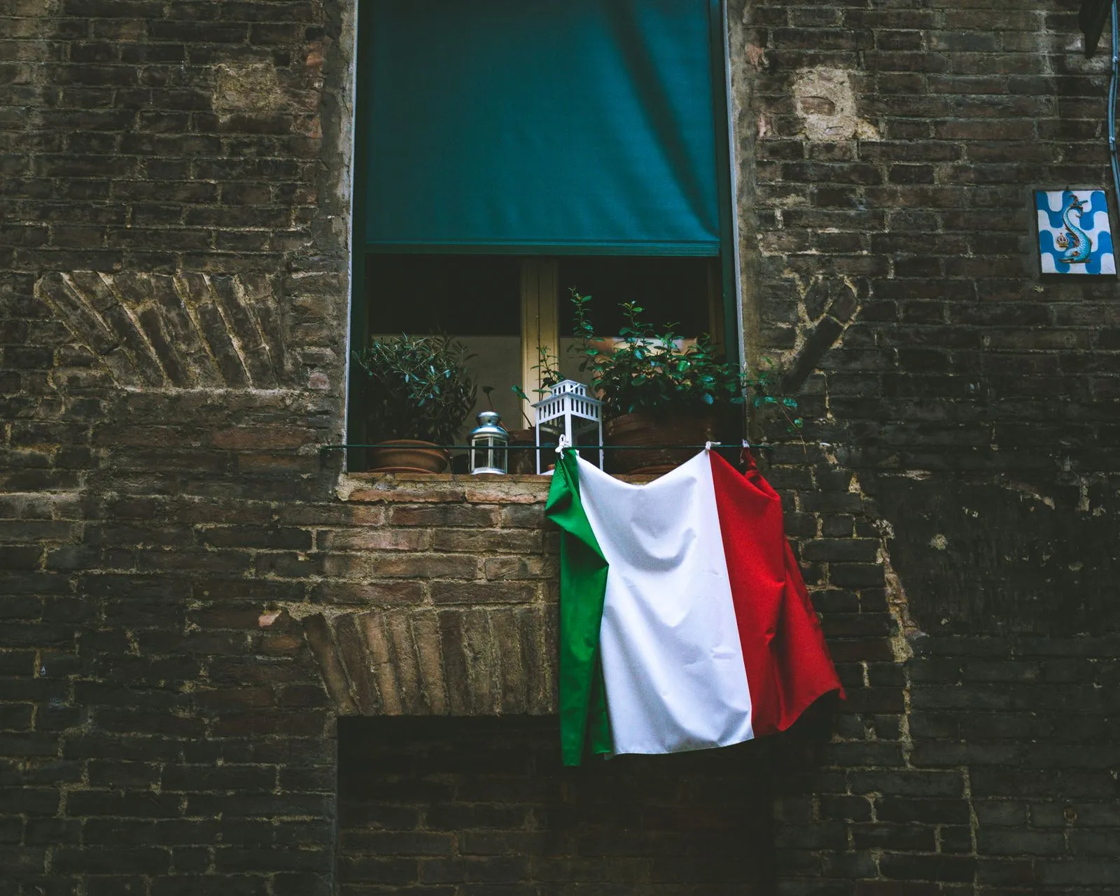 Una bandiera italiana appesa al davanzale di una finestra con piante in vaso e una lanterna, in un vecchio muro di mattoni