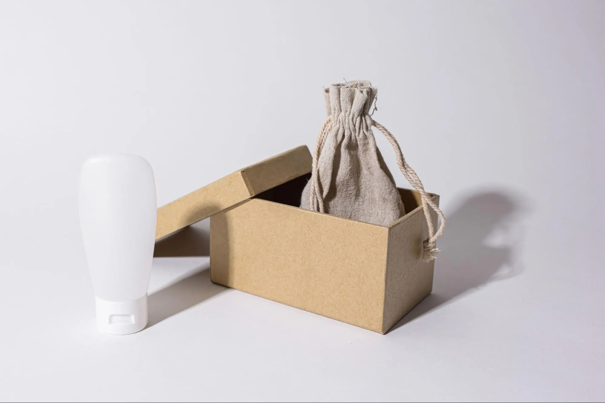 A kraft cardboard box with its lid ajar, a linen drawstring pouch inside it and a white plastic tube beside it, on a plain background