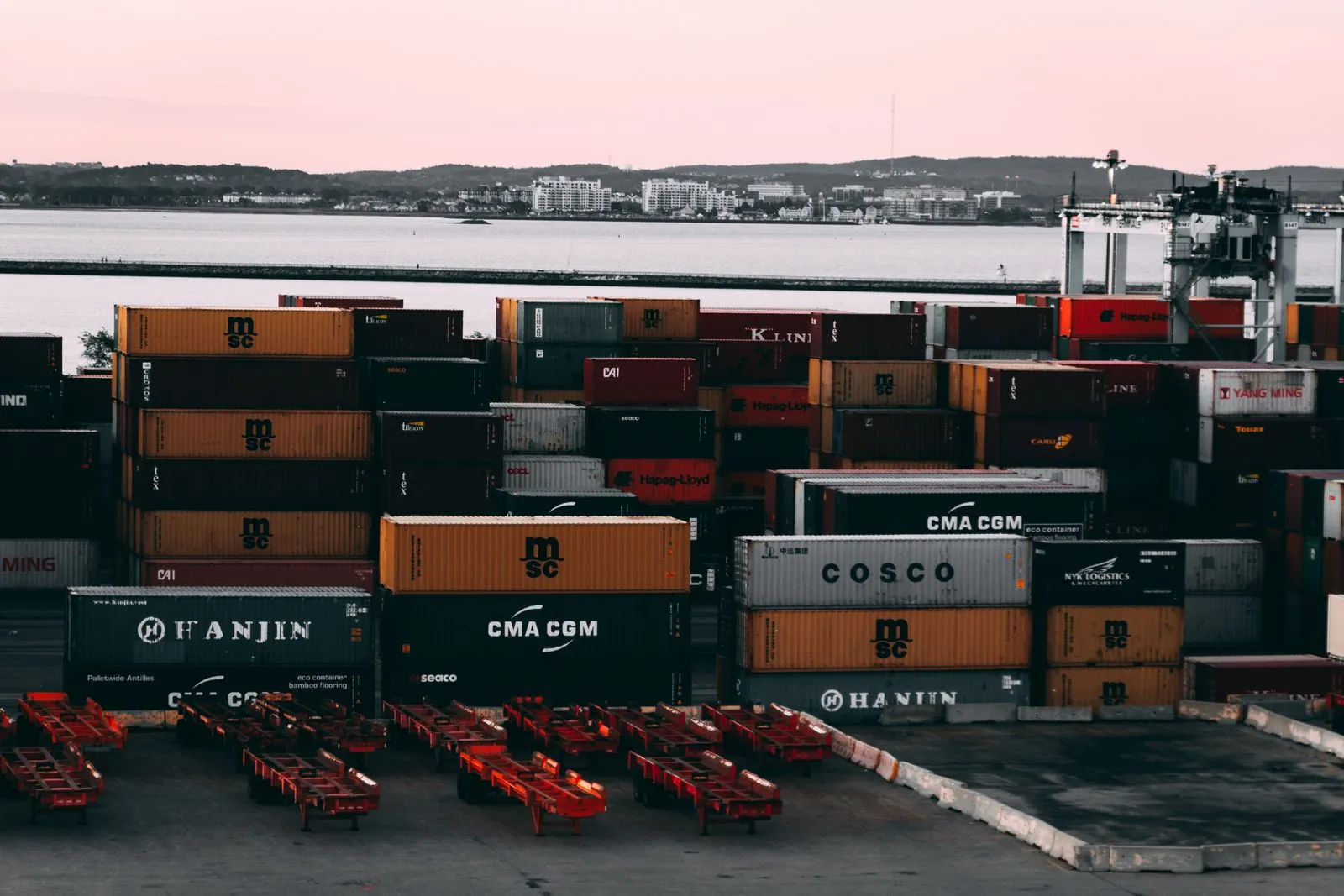 Container marittimi impilati in un porto al tramonto, con l'acqua e una città costiera sullo sfondo