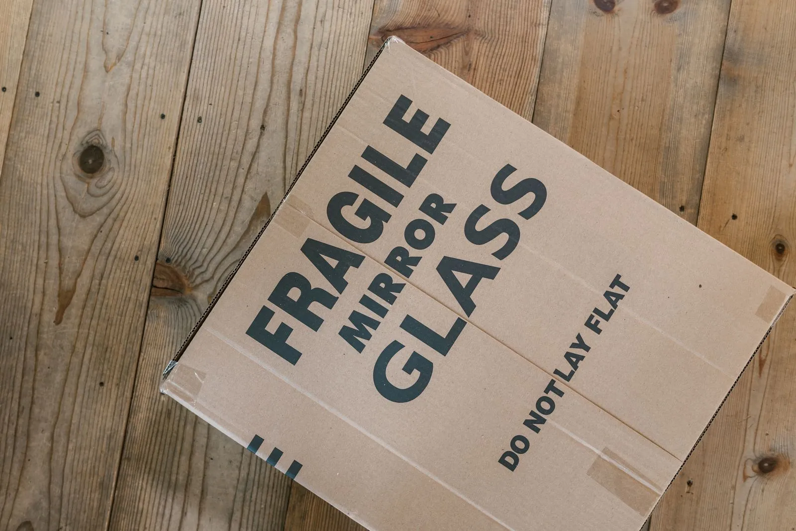 Una scatola di cartone piatta su un pavimento di legno, stampata a grandi lettere con le scritte FRAGILE, MIRROR, GLASS e DO NOT LAY FLAT