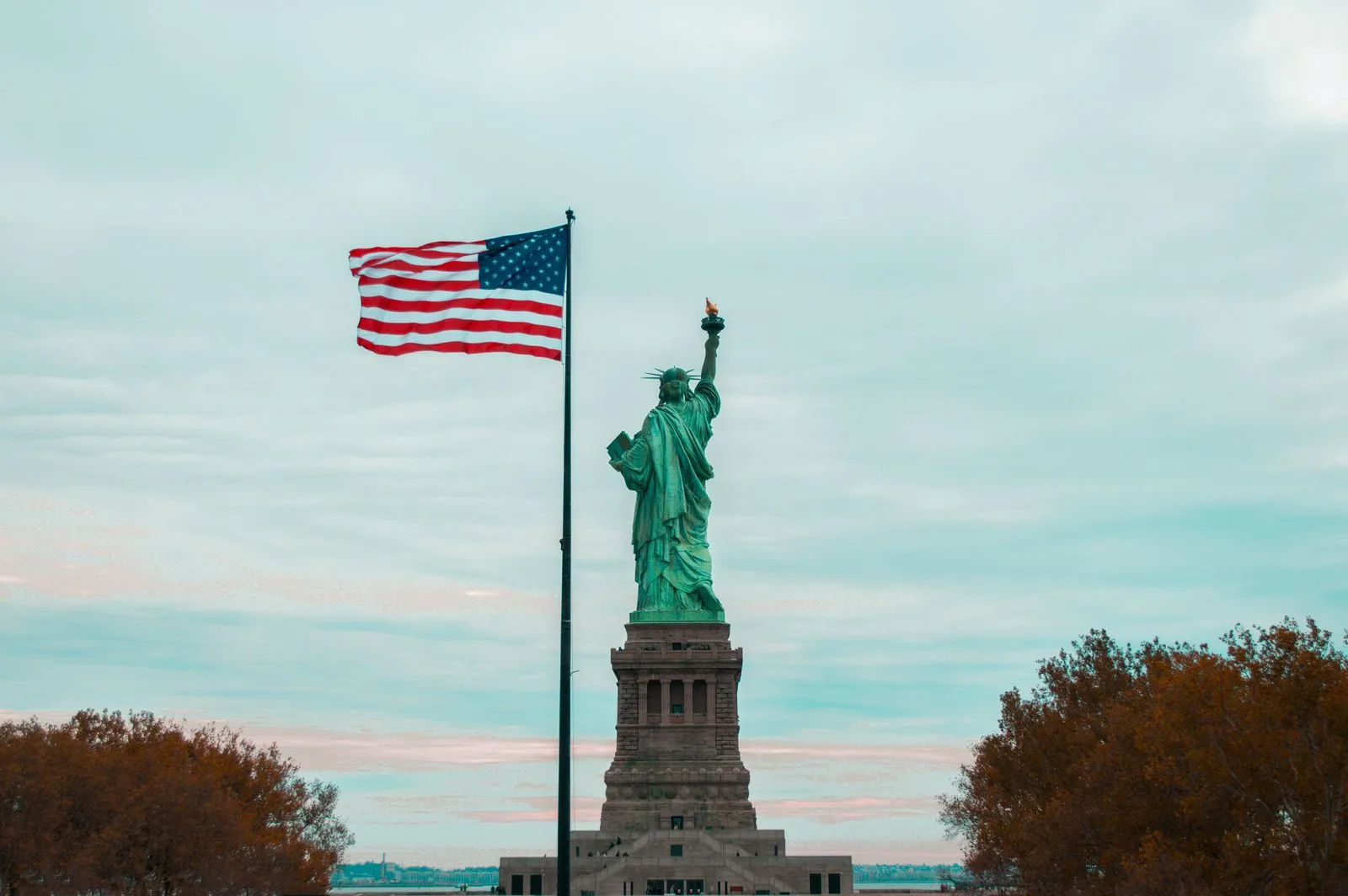 The Statue of Liberty seen from behind, next to a large American flag on a tall pole, under an overcast sky
