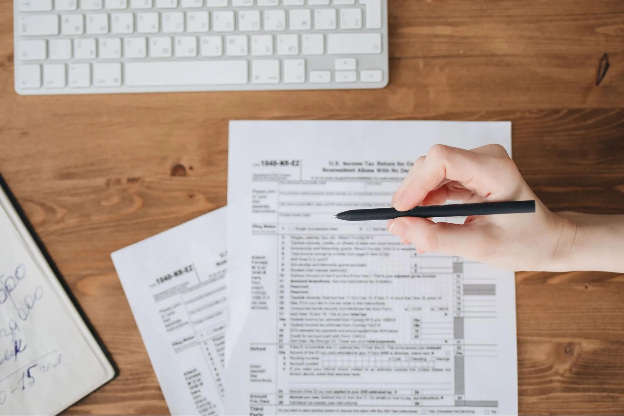 A hand filling in printed tax forms beside a keyboard on a wooden desk, representing the administrative filings behind a customs hold