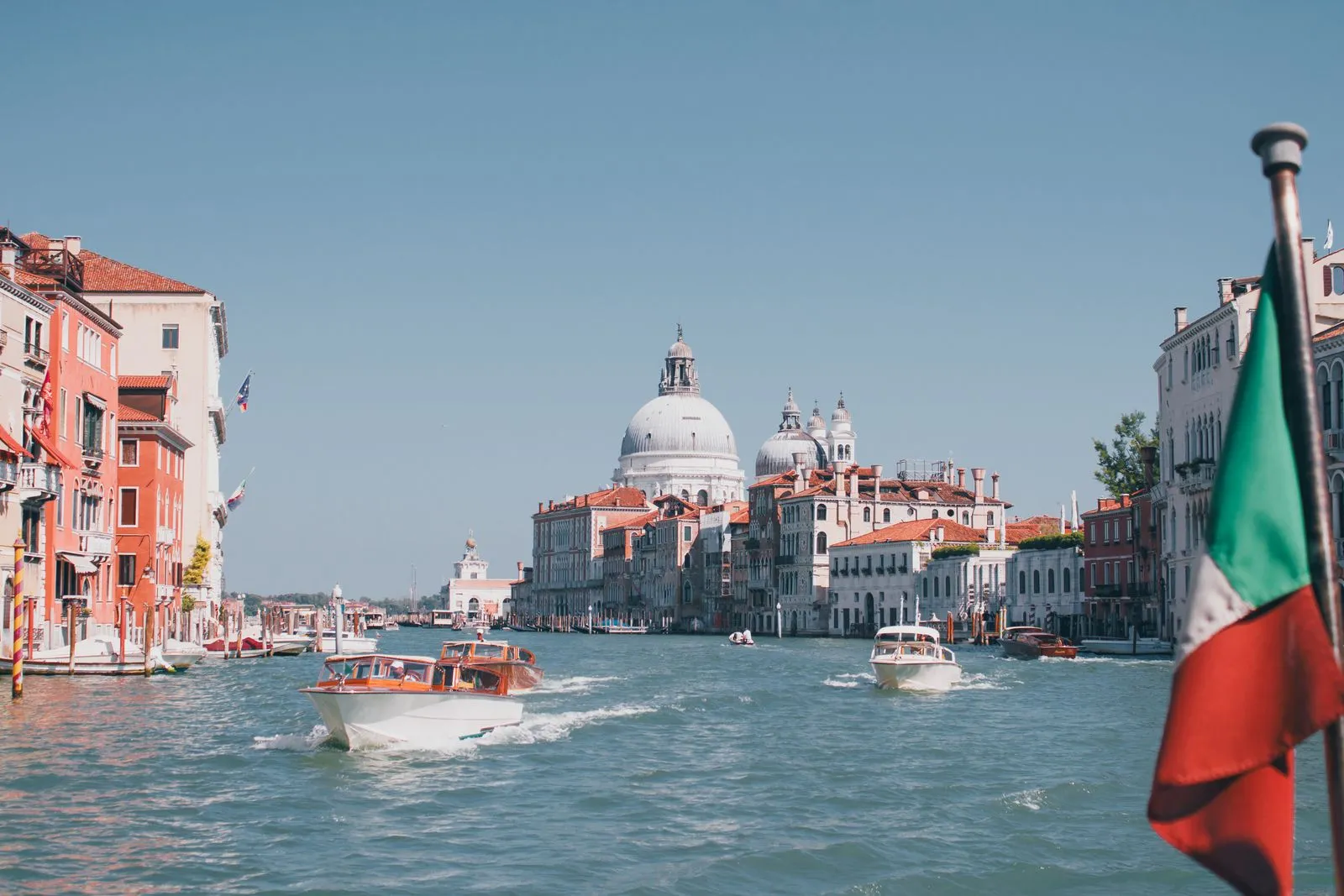 Motoscafi sul Canal Grande a Venezia diretti verso le cupole di Santa Maria della Salute, con una bandiera italiana su un'asta in primo piano