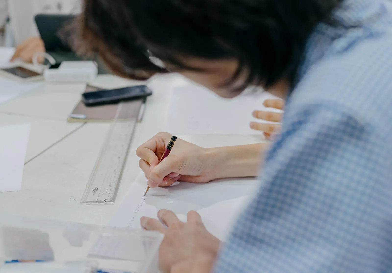 チェック柄のシャツを着た人が白い机にかがみ込み、定規の横で紙に鉛筆で書き込んでいる